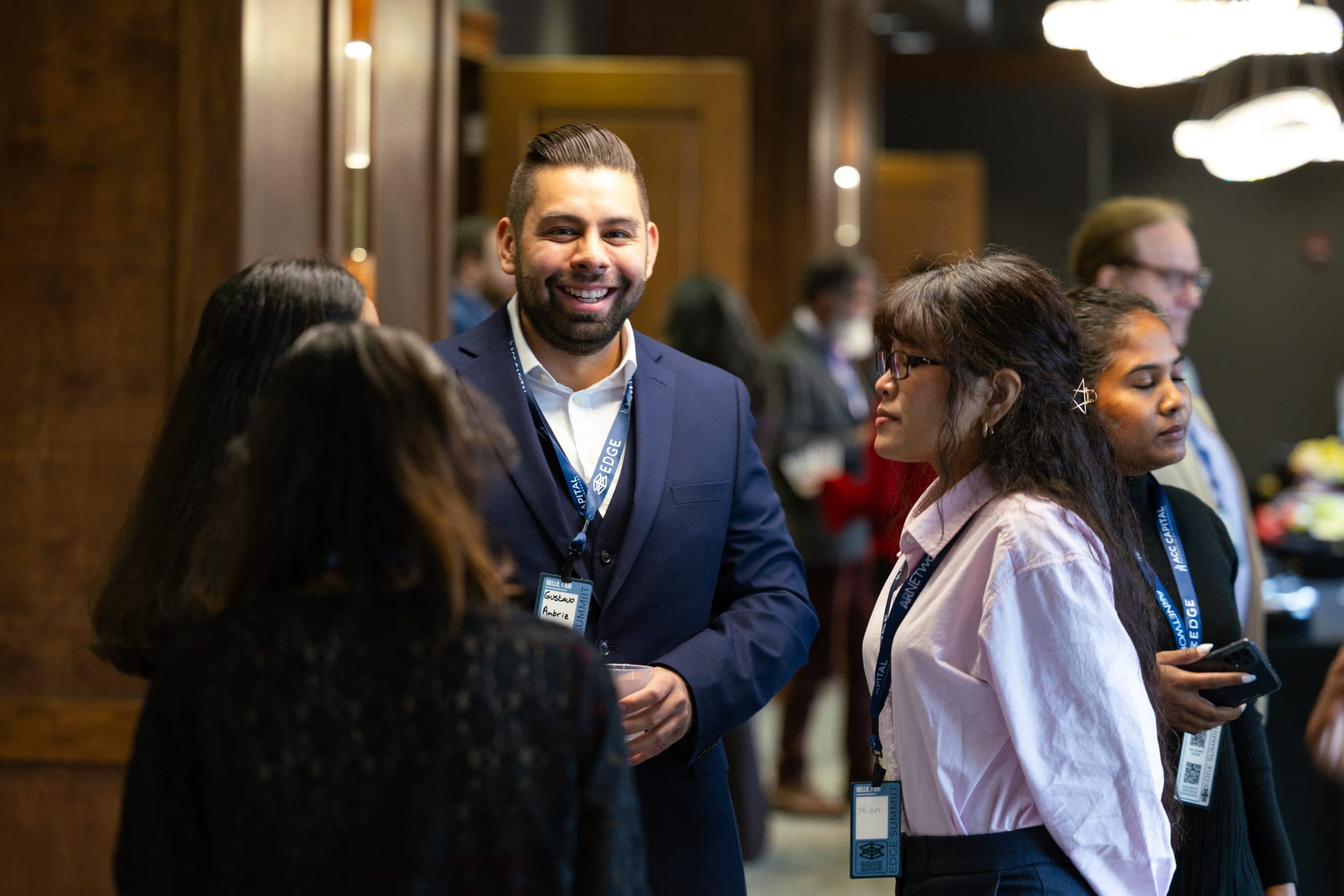 A group of people wearing conference badges converse in a lobby area. One man in a suit is smiling, and others are engaged in conversation.