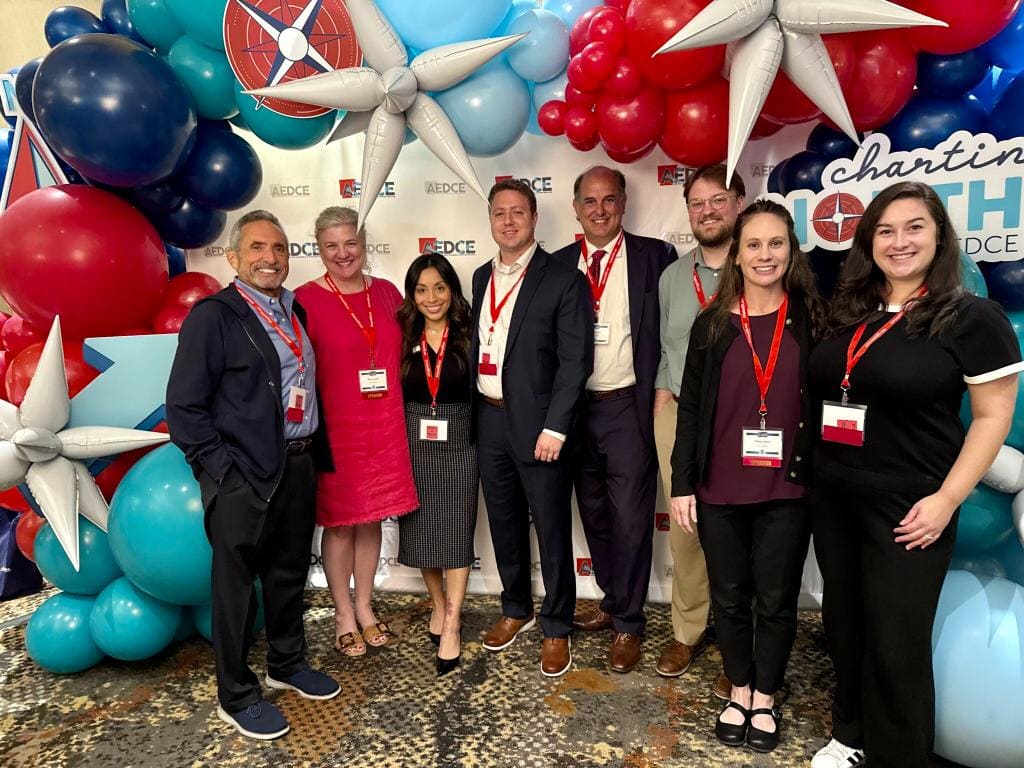 Eight adults stand together, smiling at a conference event, wearing attendee badges and red lanyards, with colorful balloon decorations and nautical-themed props in the background.