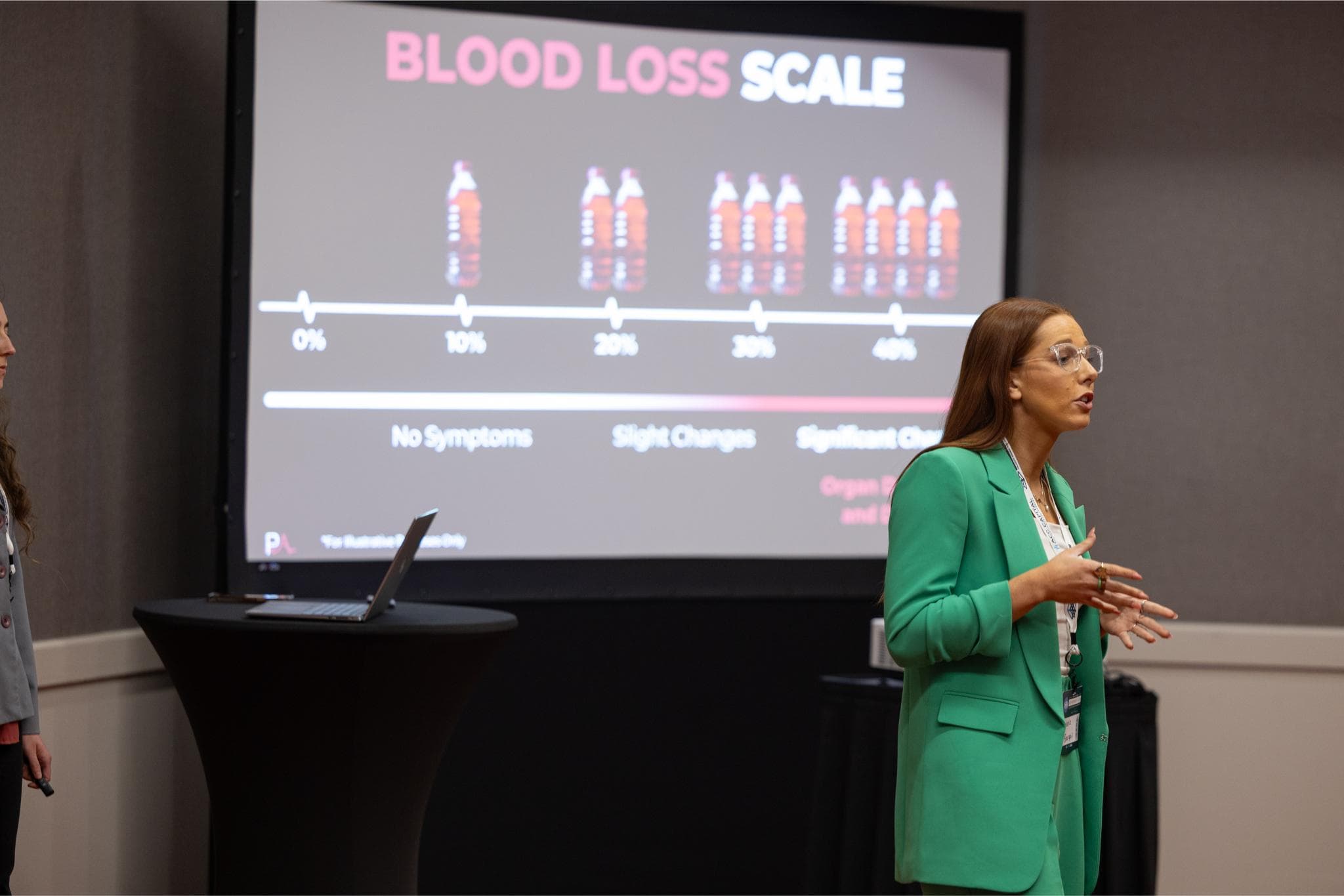 A woman in a green suit presents in front of a screen displaying a blood loss scale chart using water bottles as indicators.