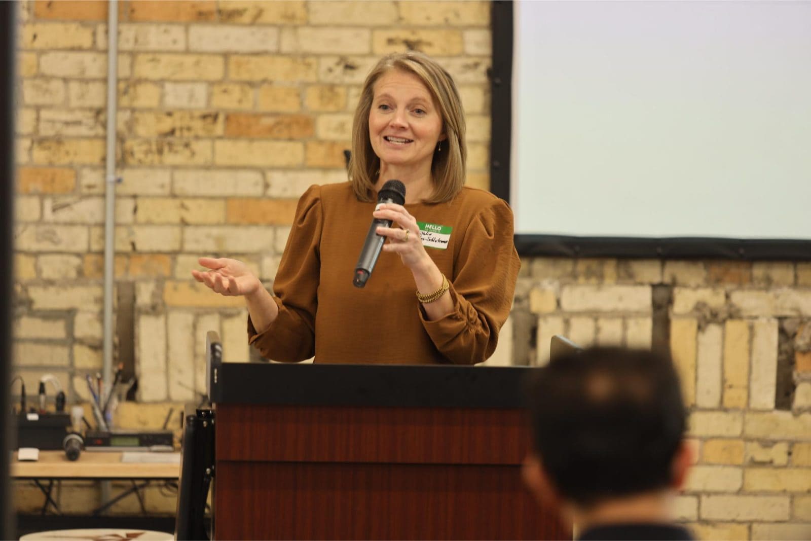 A woman stands at a podium, speaking into a microphone during an indoor event. She gestures with one hand. The background shows a brick wall and a projection screen.