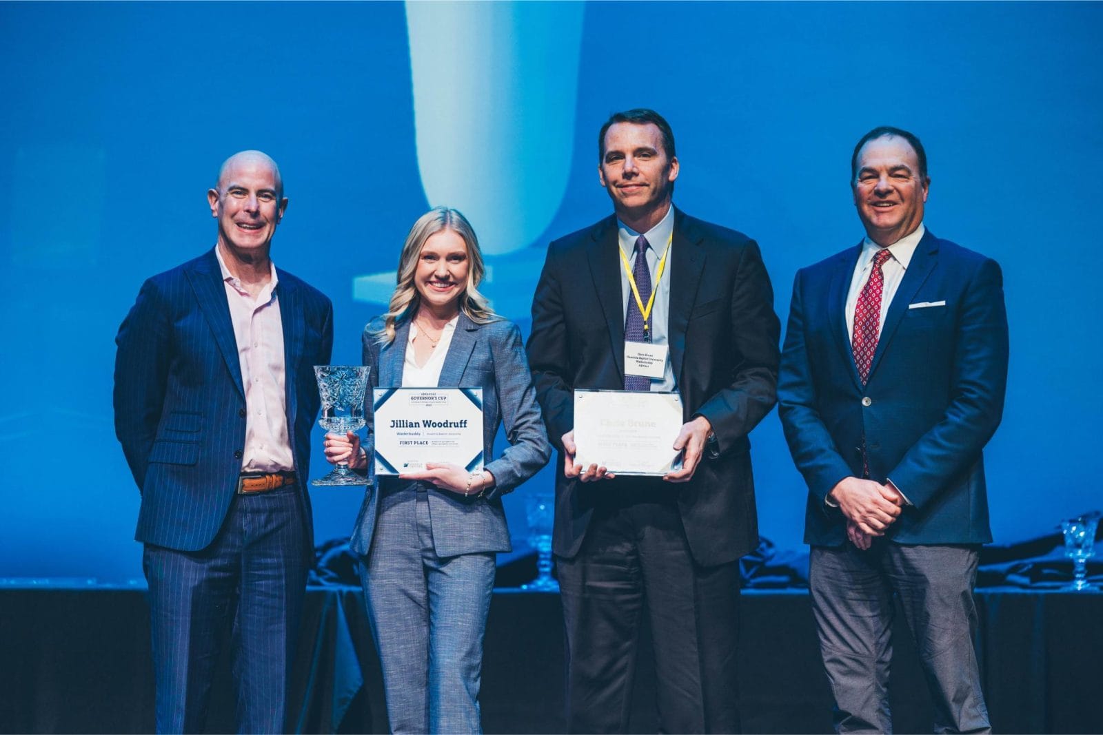 Four professionally dressed people stand on a stage; one woman holds a trophy and certificate, two men hold certificates, and all are facing the camera.