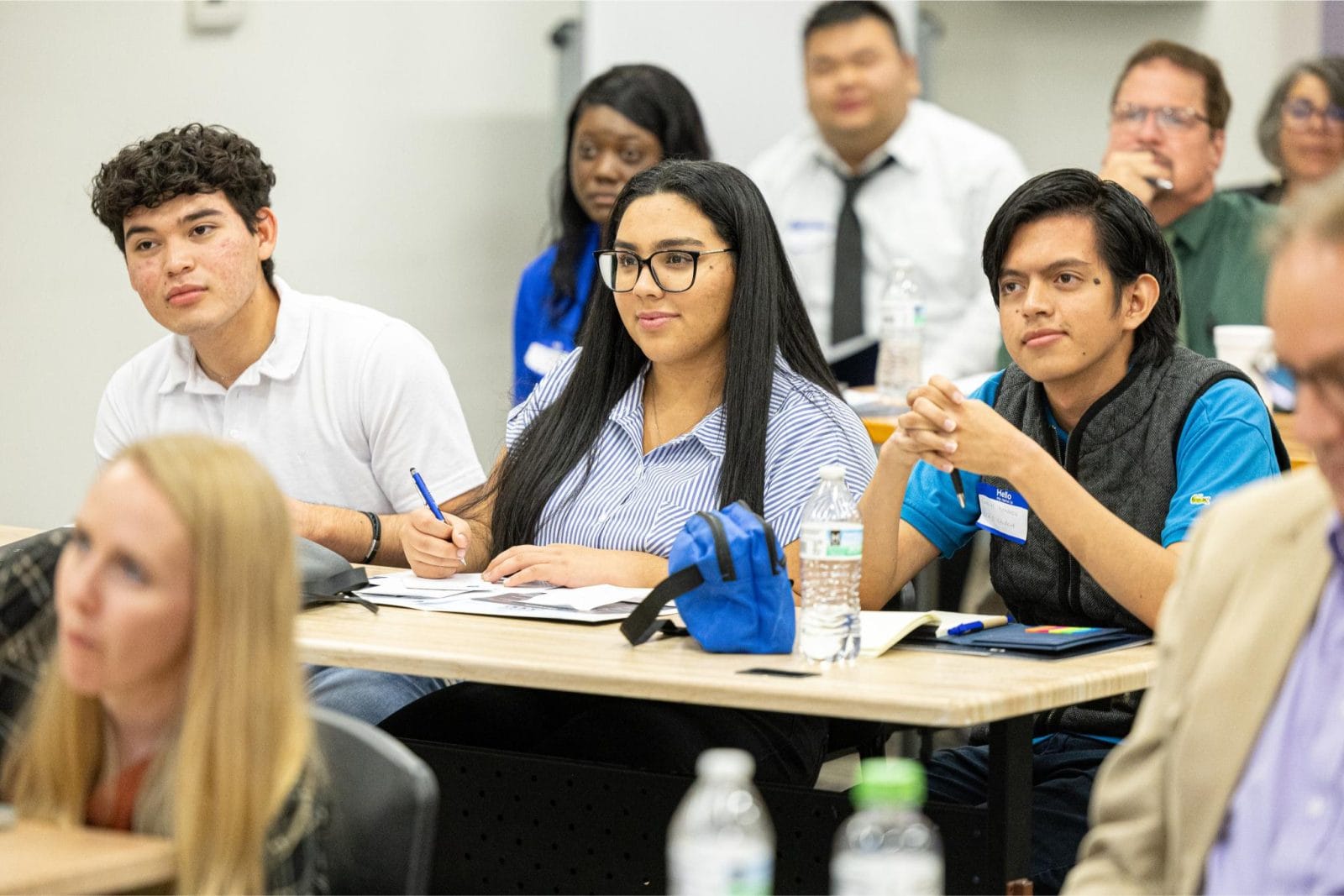 A group of students sit at desks in a classroom, listening attentively and taking notes during a lecture or seminar.