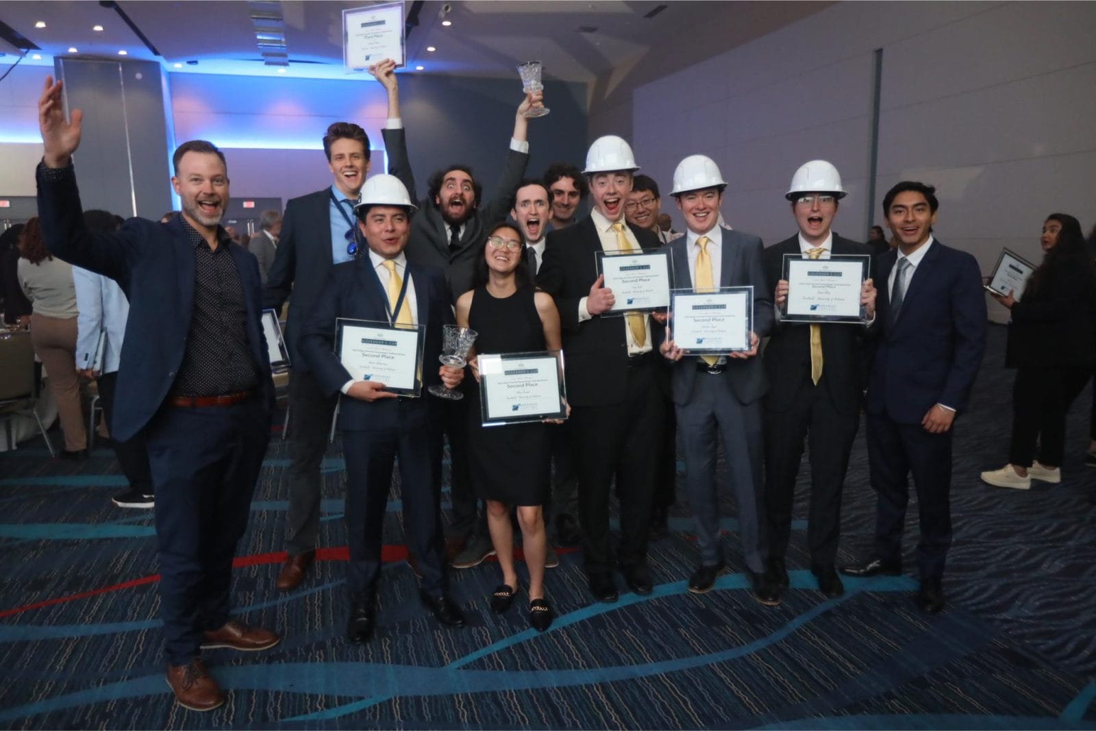 A group of people in business attire and white hard hats pose indoors, holding certificates and trophies, smiling and celebrating in a brightly lit event space.