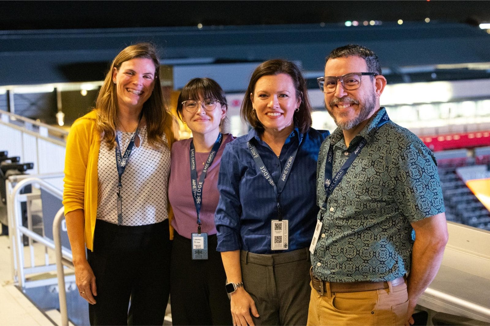 Four adults stand together smiling at an outdoor venue at night, each wearing a lanyard and name badge.
