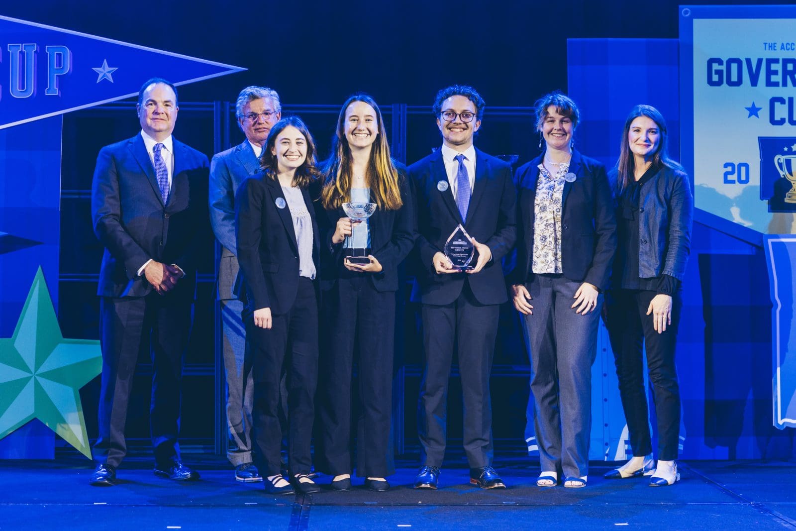 Seven people in business attire stand on a stage; two in the center hold trophies, suggesting they are the winners of an award at a formal event or competition.