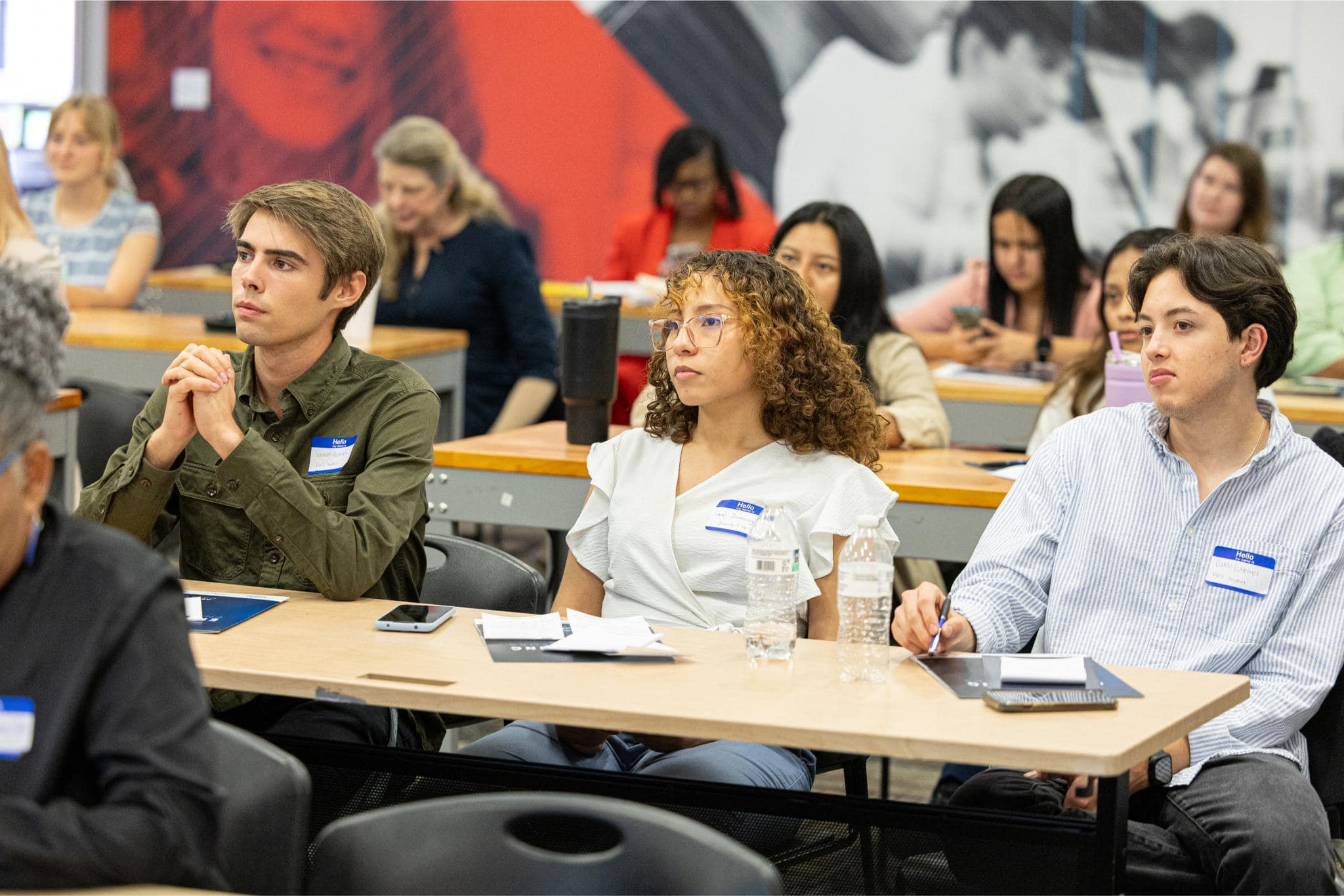A diverse group of people sit at desks in a classroom, listening attentively to a presentation. Most have notebooks or folders, and some wear nametags.