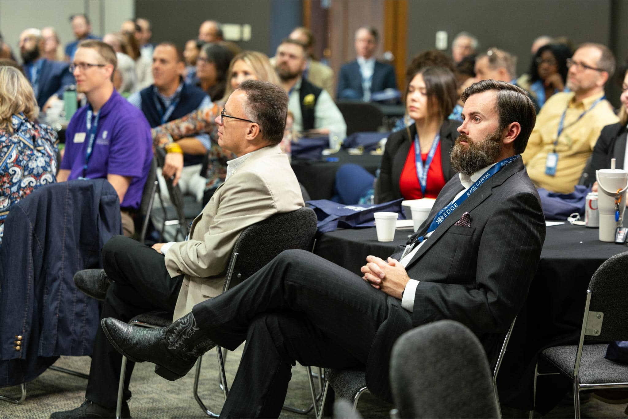 A group of people, dressed in business attire, sit at tables attentively listening during a conference or seminar in a meeting room.