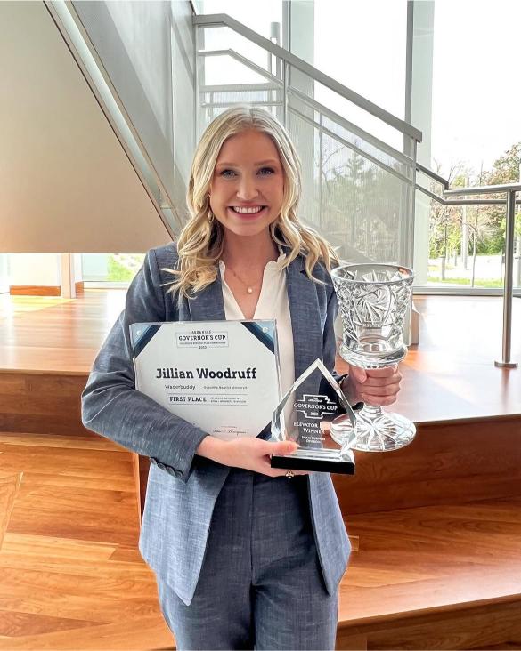 A woman in a blue suit holds a crystal trophy, a glass plaque, and a certificate that says “Jillian Woodruff, First Place,” standing indoors on a wooden floor near stairs.