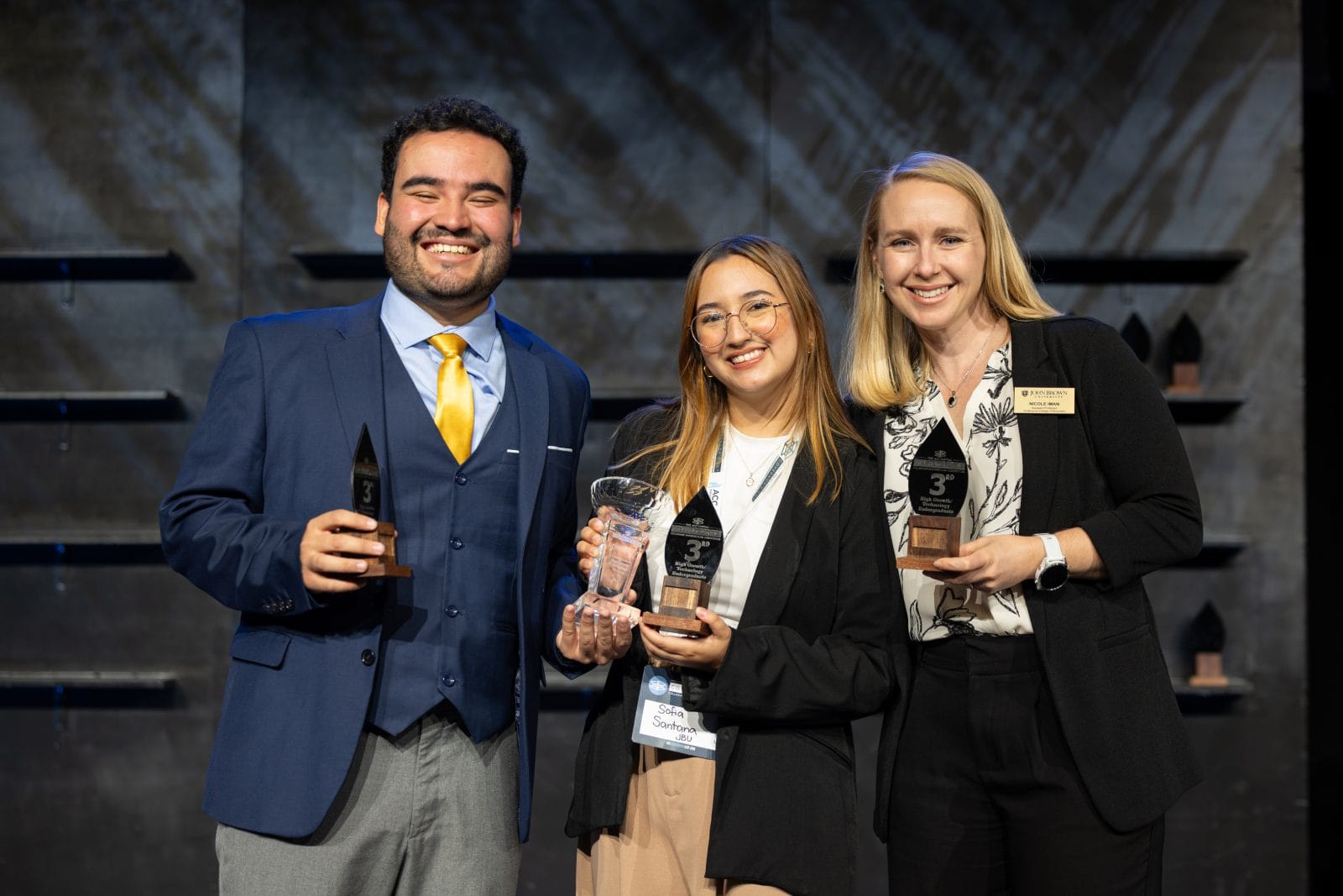 Three people in business attire stand together on stage, smiling and holding trophies and an award plaque, in front of a dark backdrop with shelves displaying more awards.