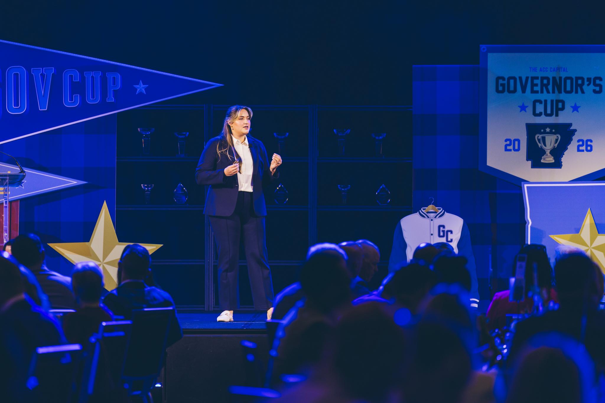 A woman in a navy suit stands on a stage addressing an audience at a Governor's Cup event, with a branded banner and varsity jacket displayed behind her.