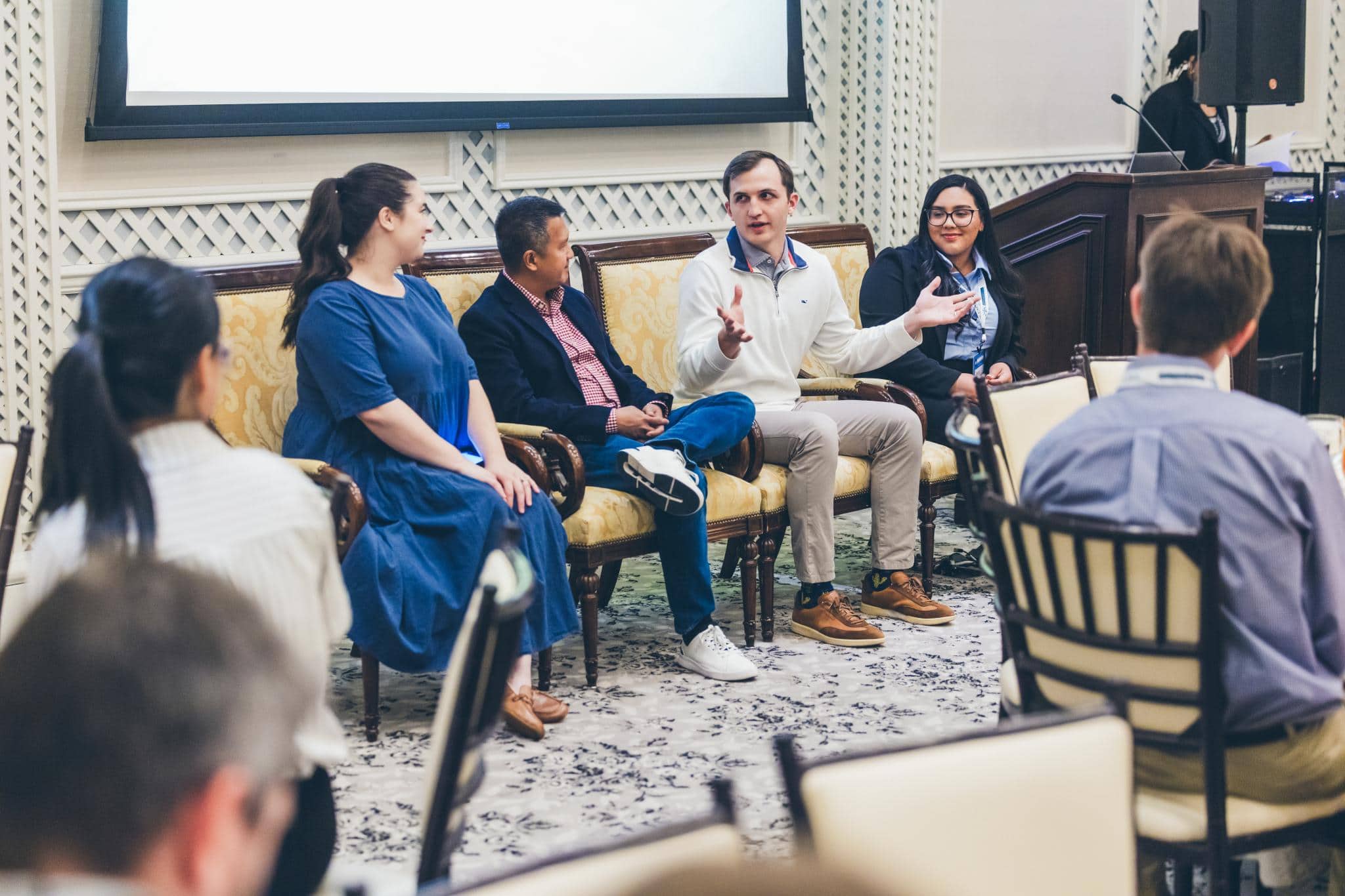 Four people sit on a couch in discussion during a panel event, with an audience seated in front of them and one person standing at a podium in the background.