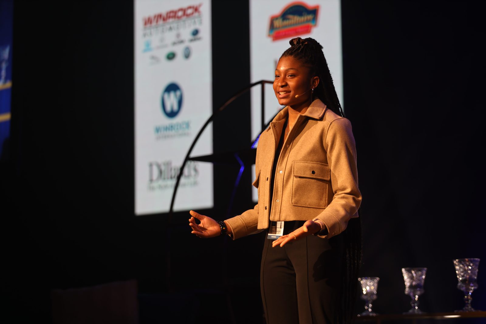 A woman stands on a stage speaking, with sponsor logos displayed on screens in the background.
