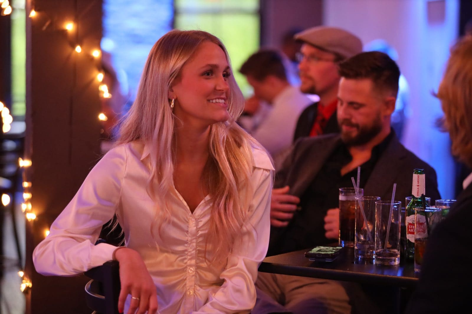 A woman with long blonde hair in a white blouse sits at a table in a dimly lit bar, smiling, with drinks and other people around her.