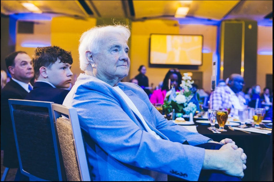 People seated at round tables in a banquet hall, listening to a presentation. The focus is on an older man in a blue blazer with others beside him. Event lighting is visible in the background.