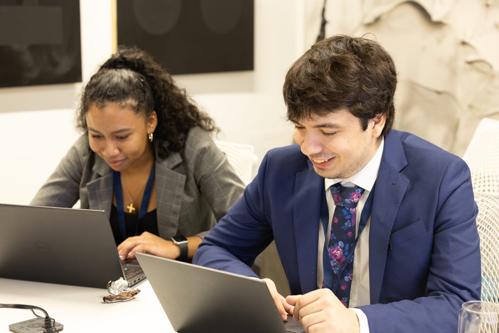 Two people sit at a table working on laptops. The woman on the left types, while the man on the right, dressed in a blue suit, smiles at his screen.