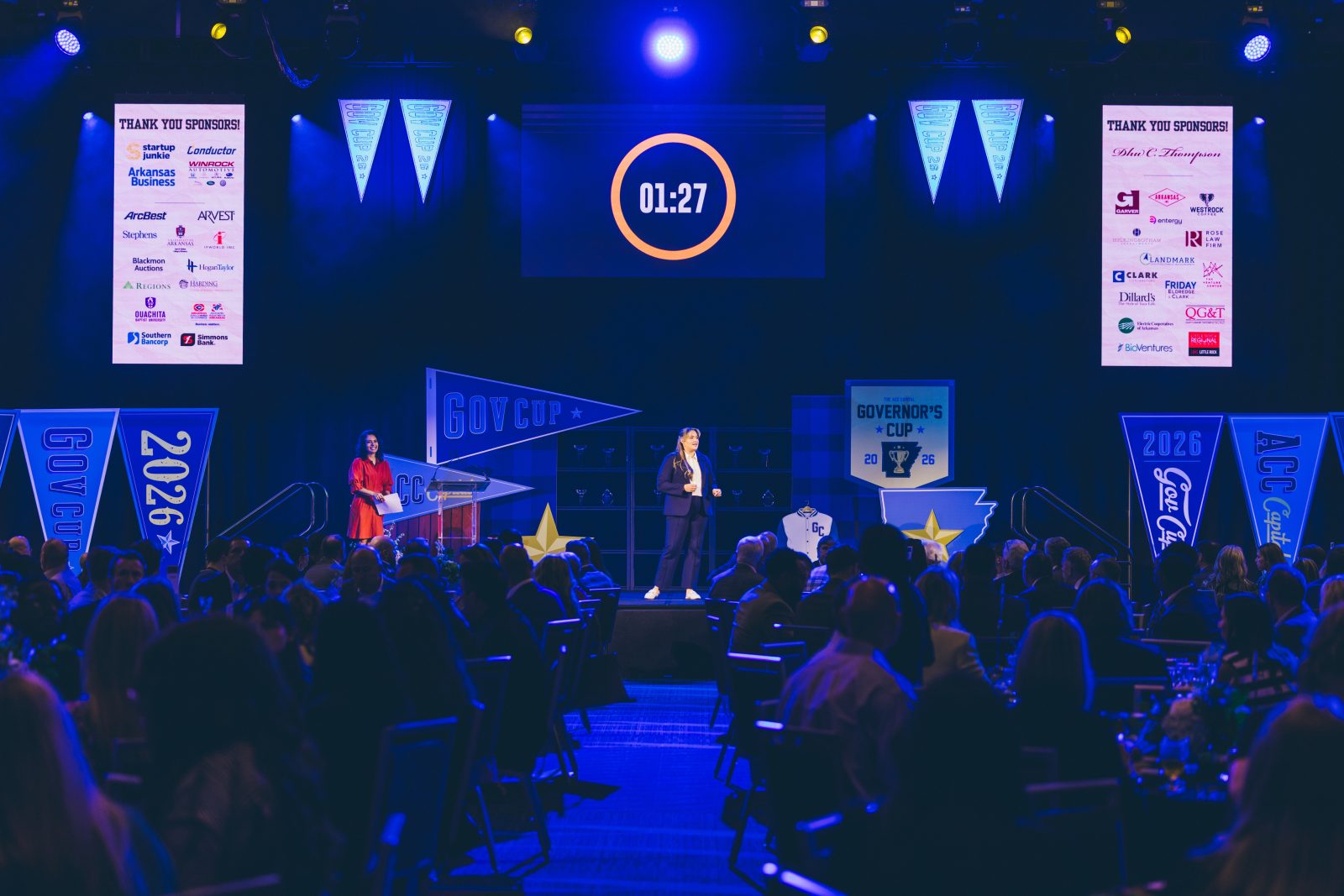 A speaker stands on stage at a formal event with a countdown timer displayed above and sponsor banners visible, while an audience watches from seated tables.