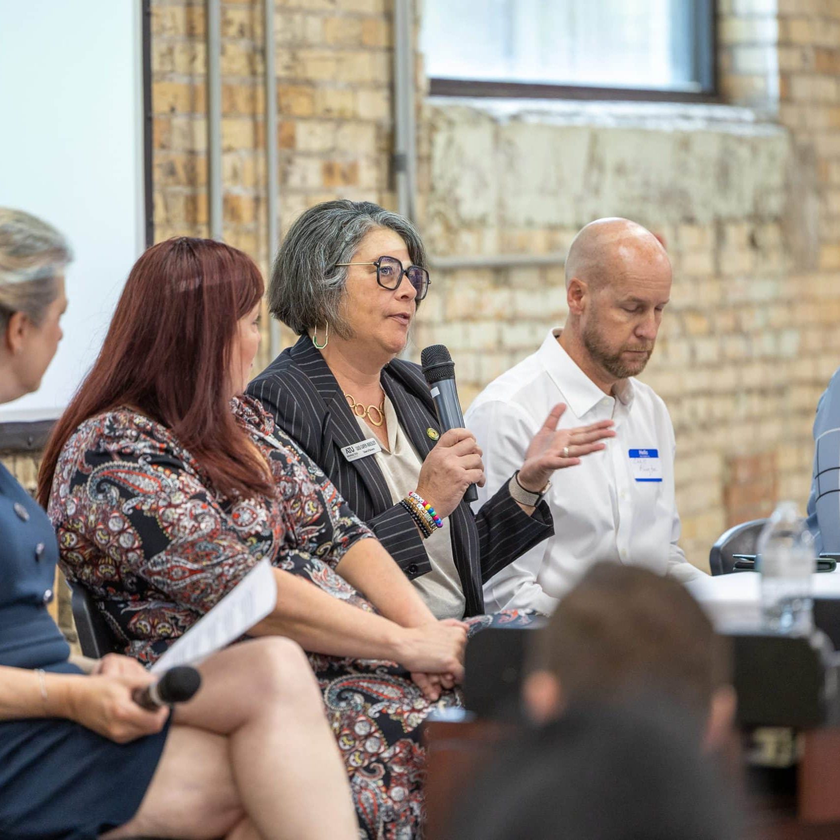 Five people sit at a table during a panel discussion; one woman speaks into a microphone while the others listen.