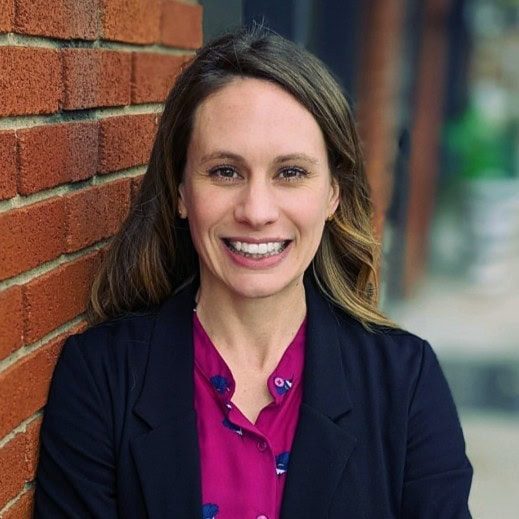 Woman with brown hair, wearing a dark blazer and magenta blouse, smiles while standing against a brick wall with an outdoor blurred background.