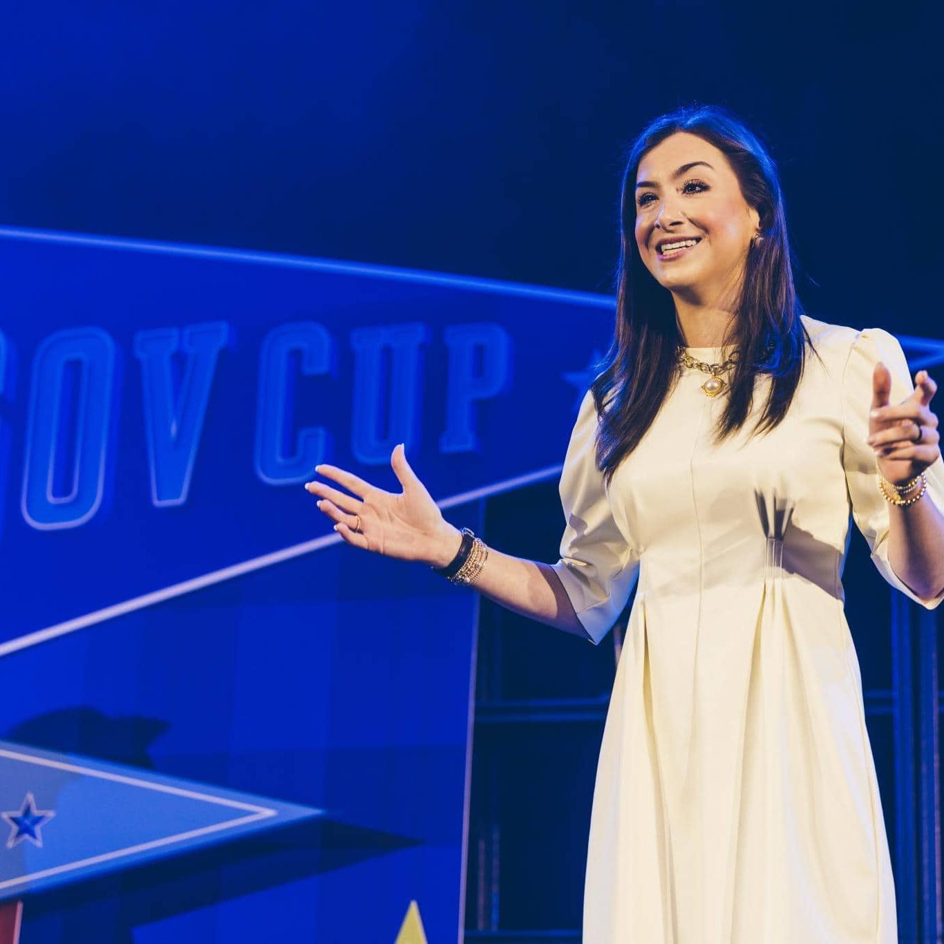A woman in a white dress speaks on stage with arms open, standing in front of a blue "GOV CUP" sign and a display shelf.