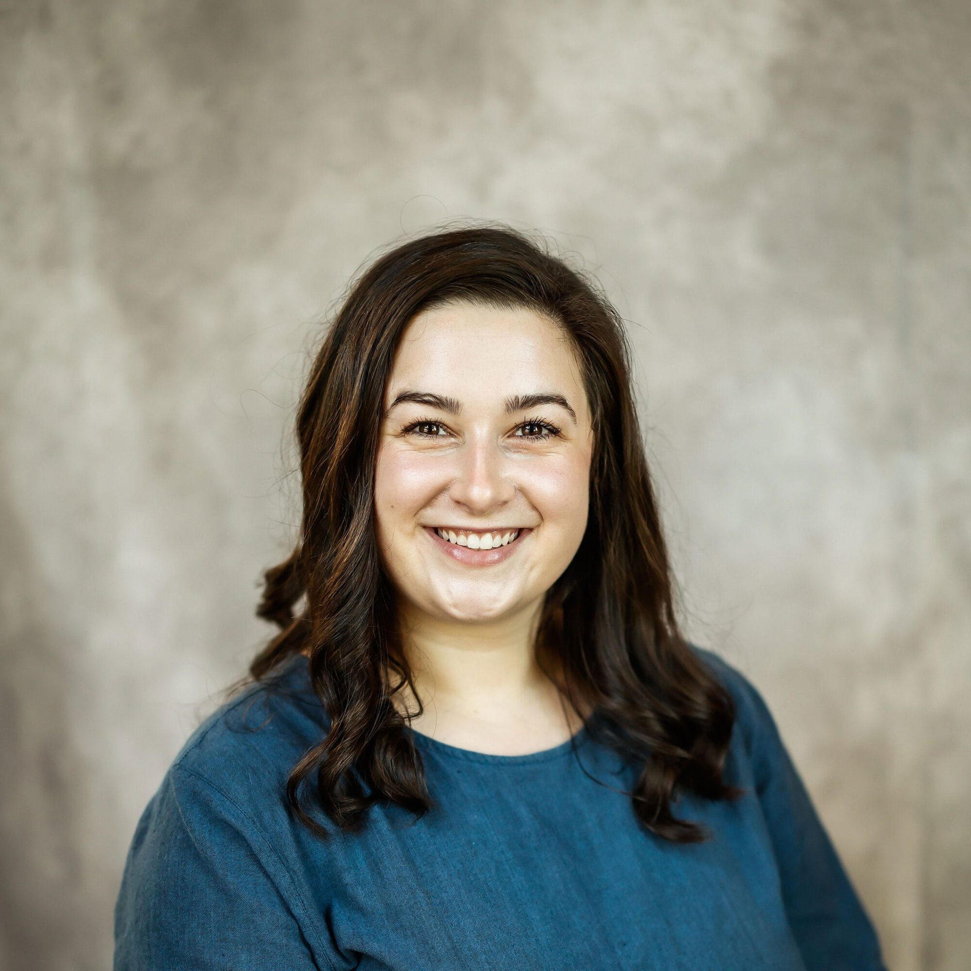 A woman with long brown hair, wearing a blue top, smiles at the camera with a neutral beige background behind her.