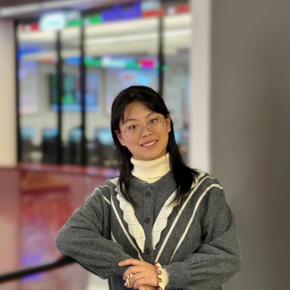 A person wearing glasses and a gray sweater stands indoors, smiling with arms crossed in front of them. Blurred office space with screens is visible in the background.