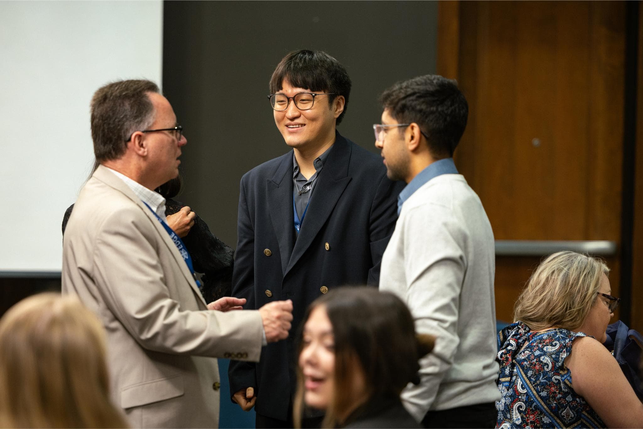 Three men standing and talking at an indoor event, with other people seated and conversing around them.