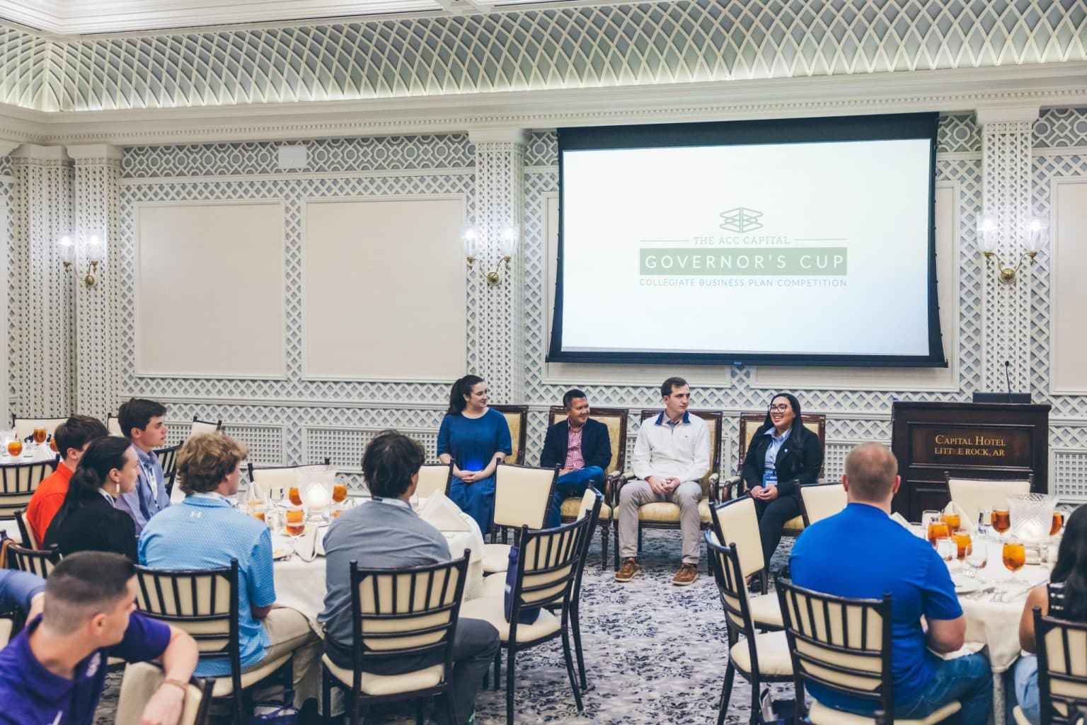 A panel of five people speaks to an audience at round tables during the Governor’s Cup Collegiate Business Plan Competition at the Capitol Hotel.