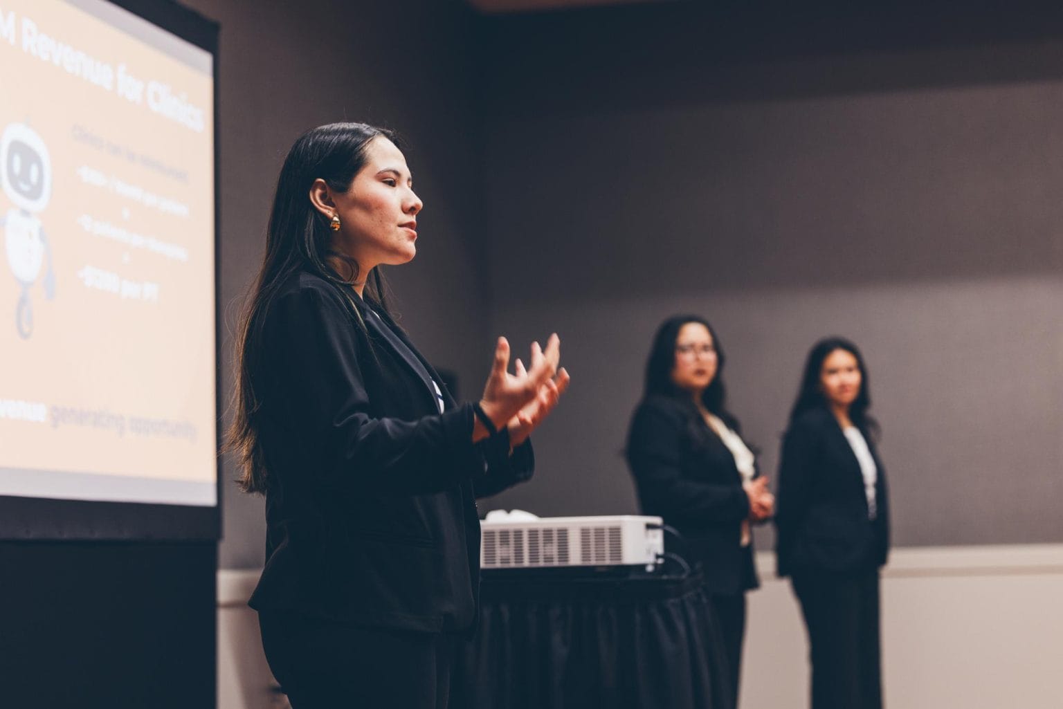 A woman in a black blazer gestures while presenting to an audience, with two other women in business attire standing in the background. A screen showing business revenue projections is on the left.