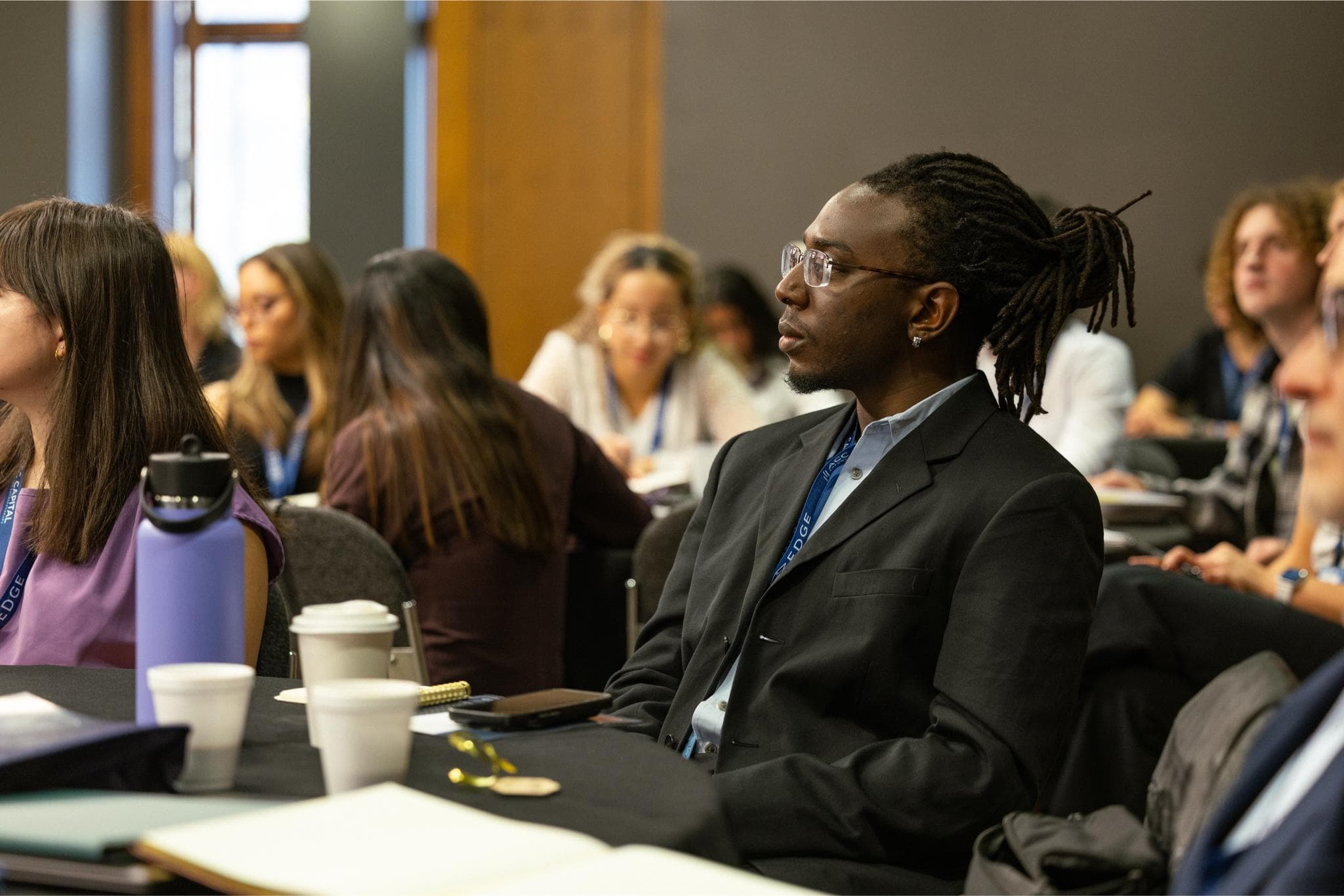 People sit at tables in a conference room, listening attentively. The foreground shows a man in a suit and glasses with a notebook, coffee cups, and a water bottle on the table.