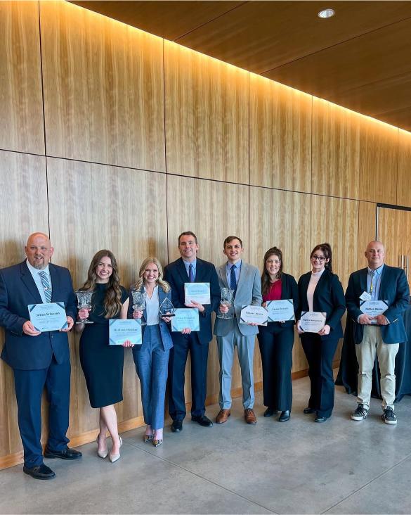Seven people in business attire stand indoors holding certificates and glass awards, posing for a group photo against a wooden wall.