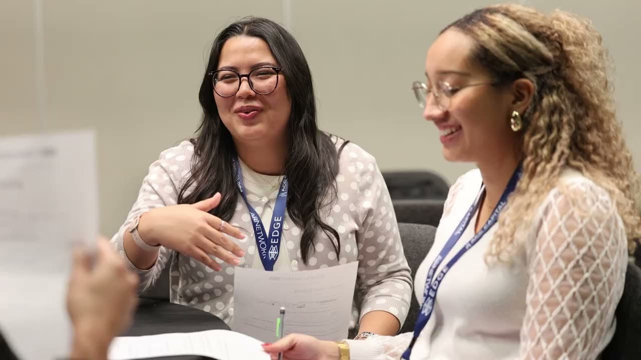 Two women sit at a table smiling and holding papers, engaged in conversation during a meeting or workshop.