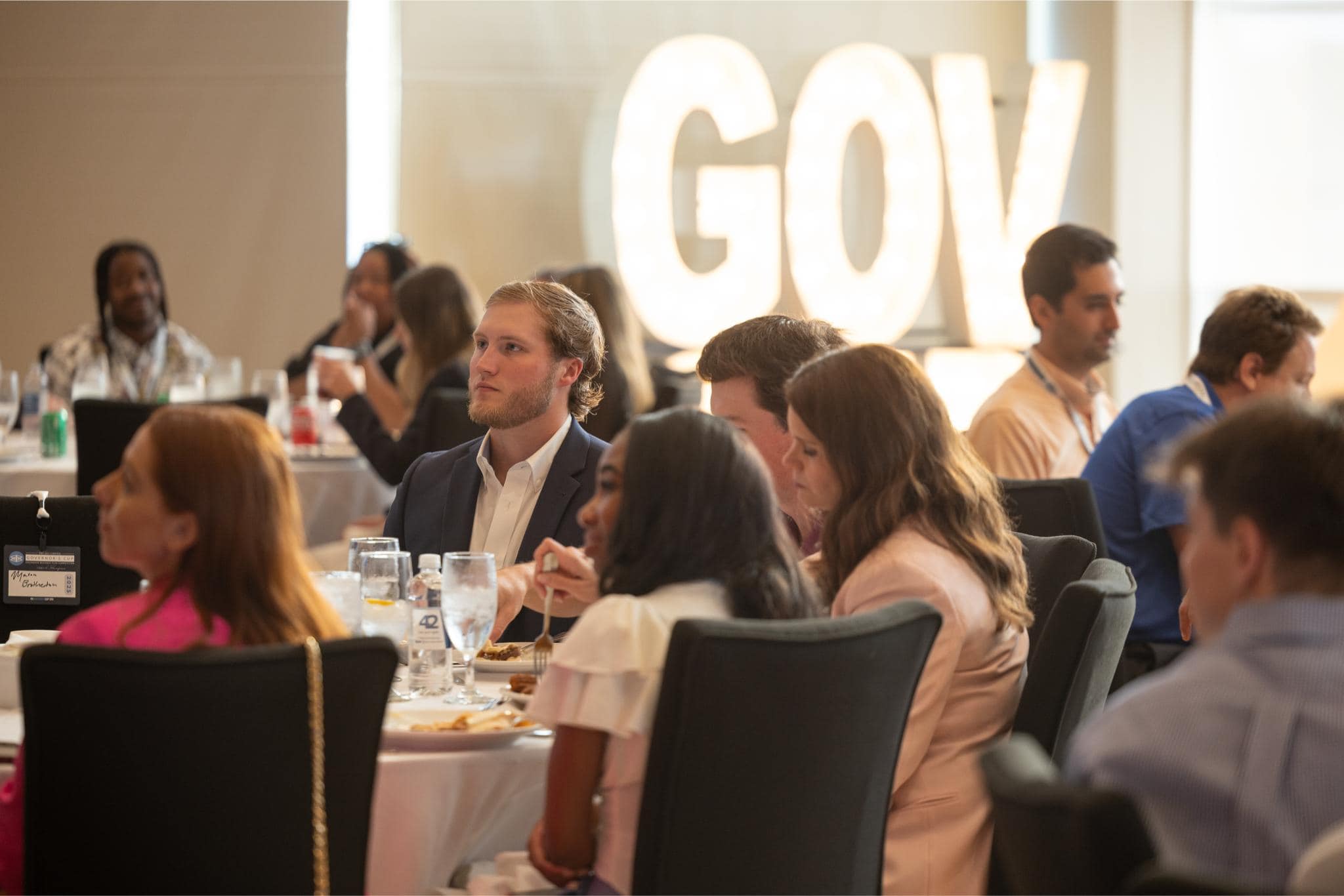 People sit at round tables in a conference room, listening attentively. Large illuminated letters in the background spell "GOV." Plates, drinks, and papers are on the tables.