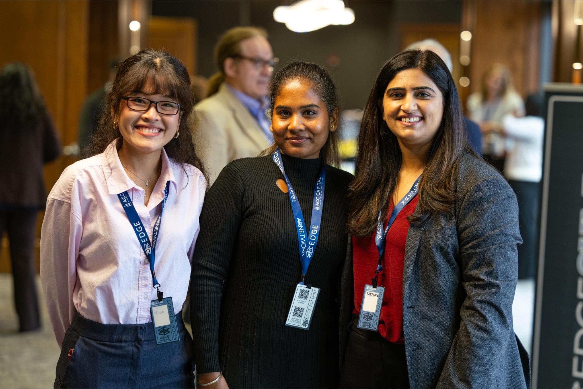Three women wearing conference badges and business attire pose together and smile for a photo indoors at a professional event.