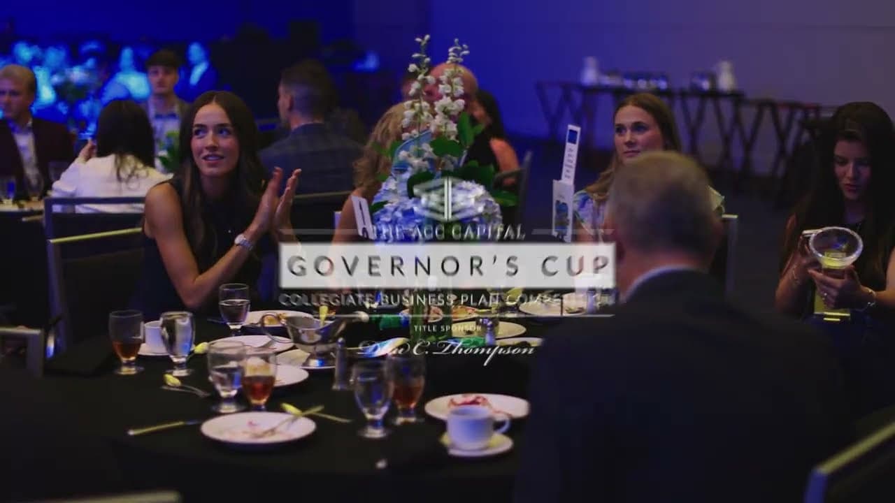 People sit at round banquet tables with empty plates and drinks at a formal event, with a “Governor’s Cup” sign visible in the foreground.